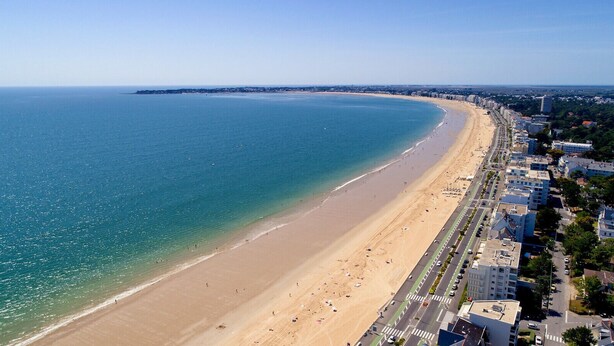 Un paysage urbain côtier avec une vaste plage de sable, une mer bleue et limpide, et une rangée de bâtiments le long de la côte.