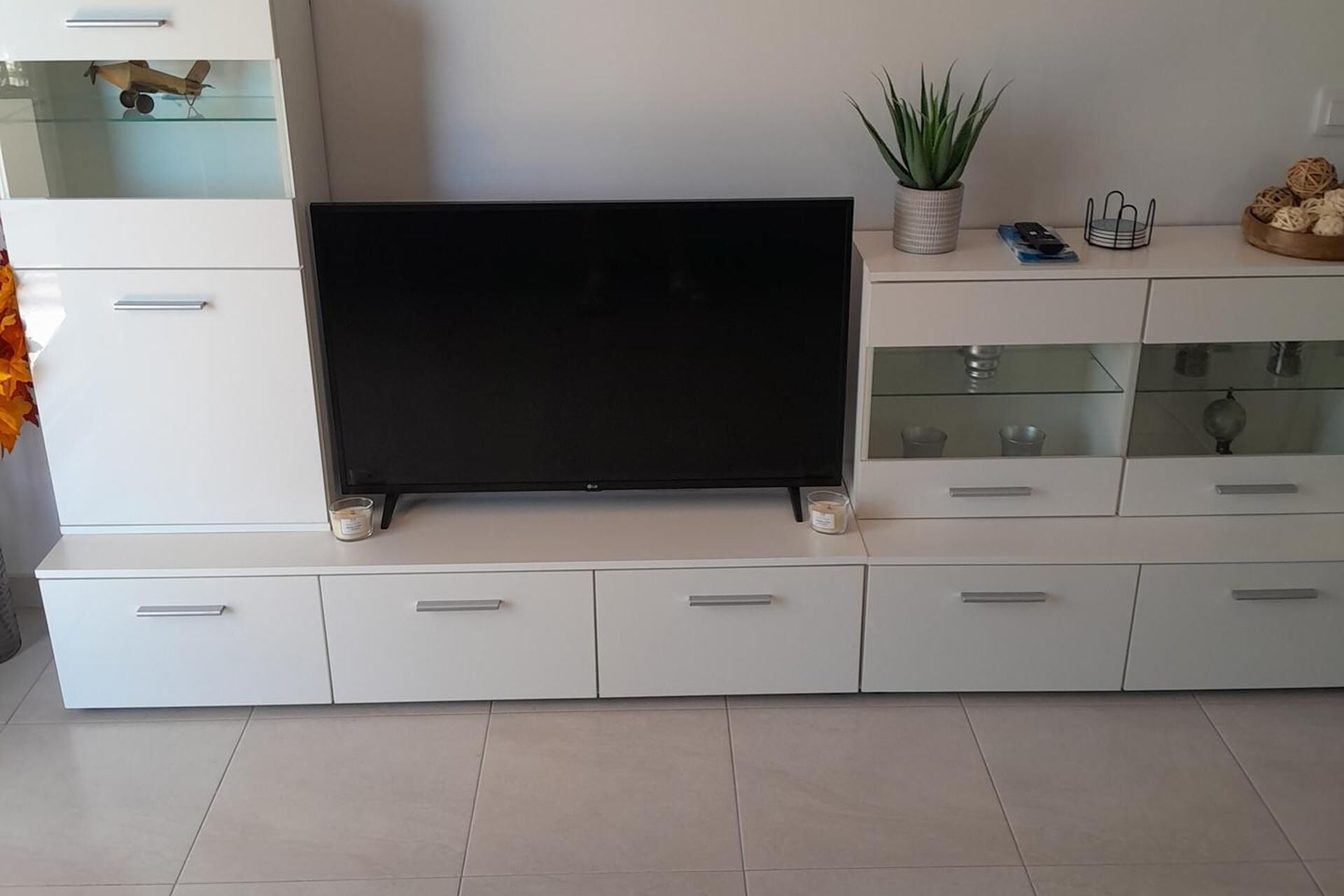 Ett modernt vardagsrum med en platt-TV placerad på en vit TV-bänk, en krukväxt och ett litet bord med dekorativa föremål.