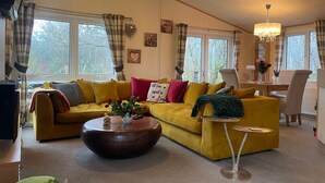A cozy living room with a yellow sofa, a round wooden coffee table, and a dining area with a chair and table. - Lilac Lodge Retreat for 6, Set in One Acre of Land (Otley)