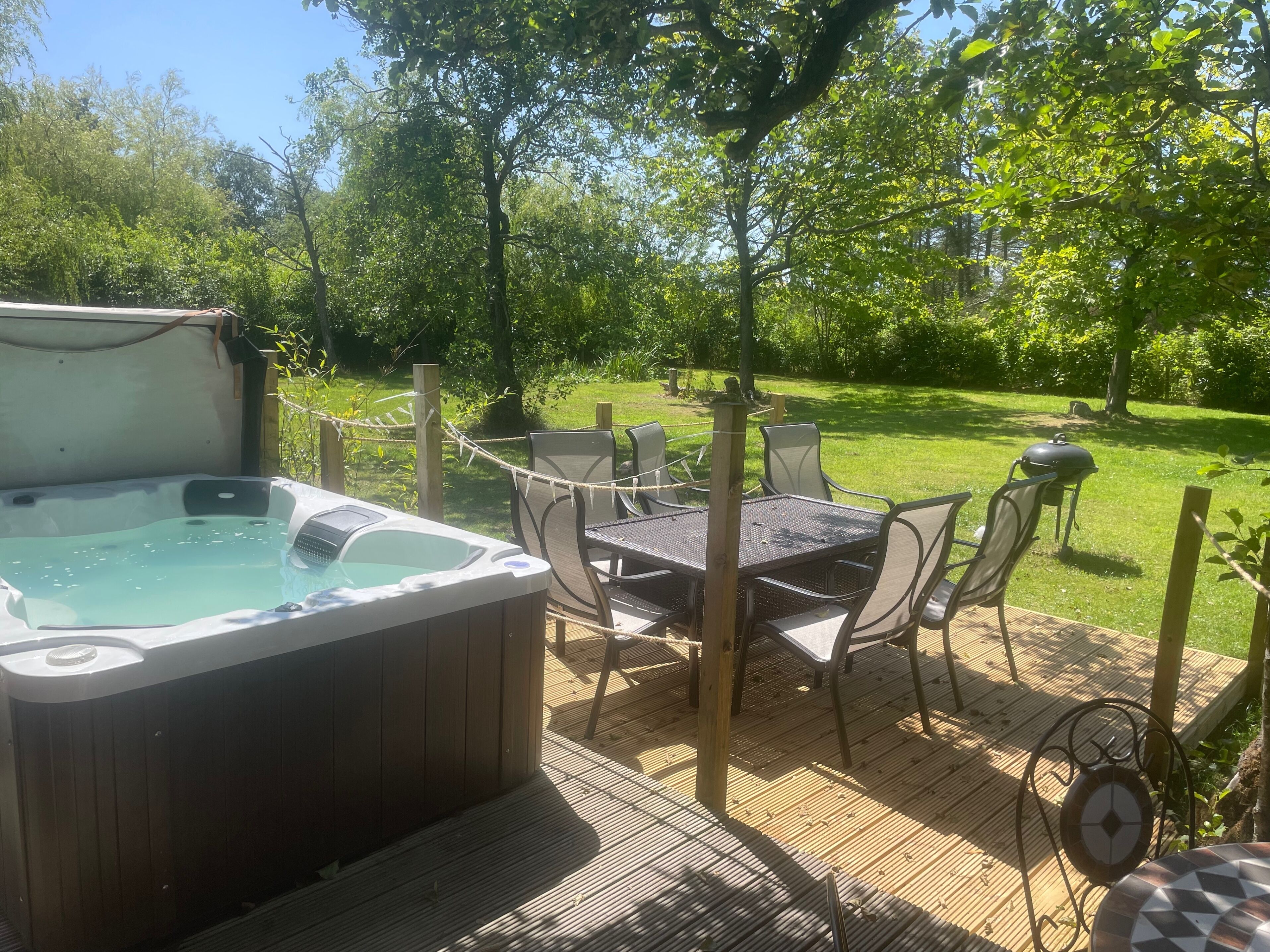 A hot tub on a wooden deck with outdoor furniture, surrounded by a rope fence and greenery.