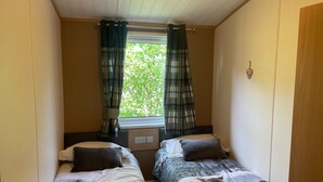 A small room with two beds, a window with curtains, and a wall-mounted light. - Lilac Lodge Retreat for 6, Set in One Acre of Land (Otley)