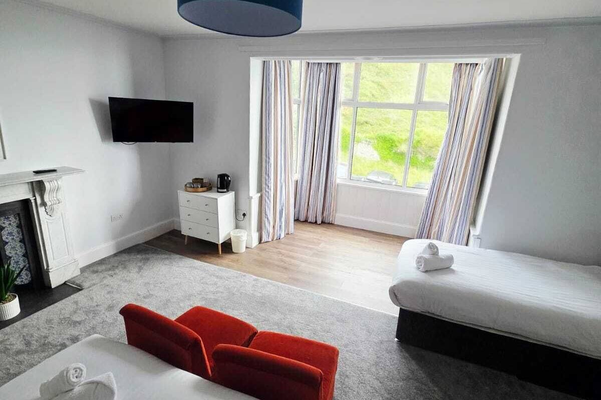 Ein modernes Hotelzimmer mit einem großen Bett, einem Einzelbett, einem roten Sofa, einem Flachbildfernseher, einem Schreibtisch mit Kaffeemaschine und einem Fenster mit Vorhängen.