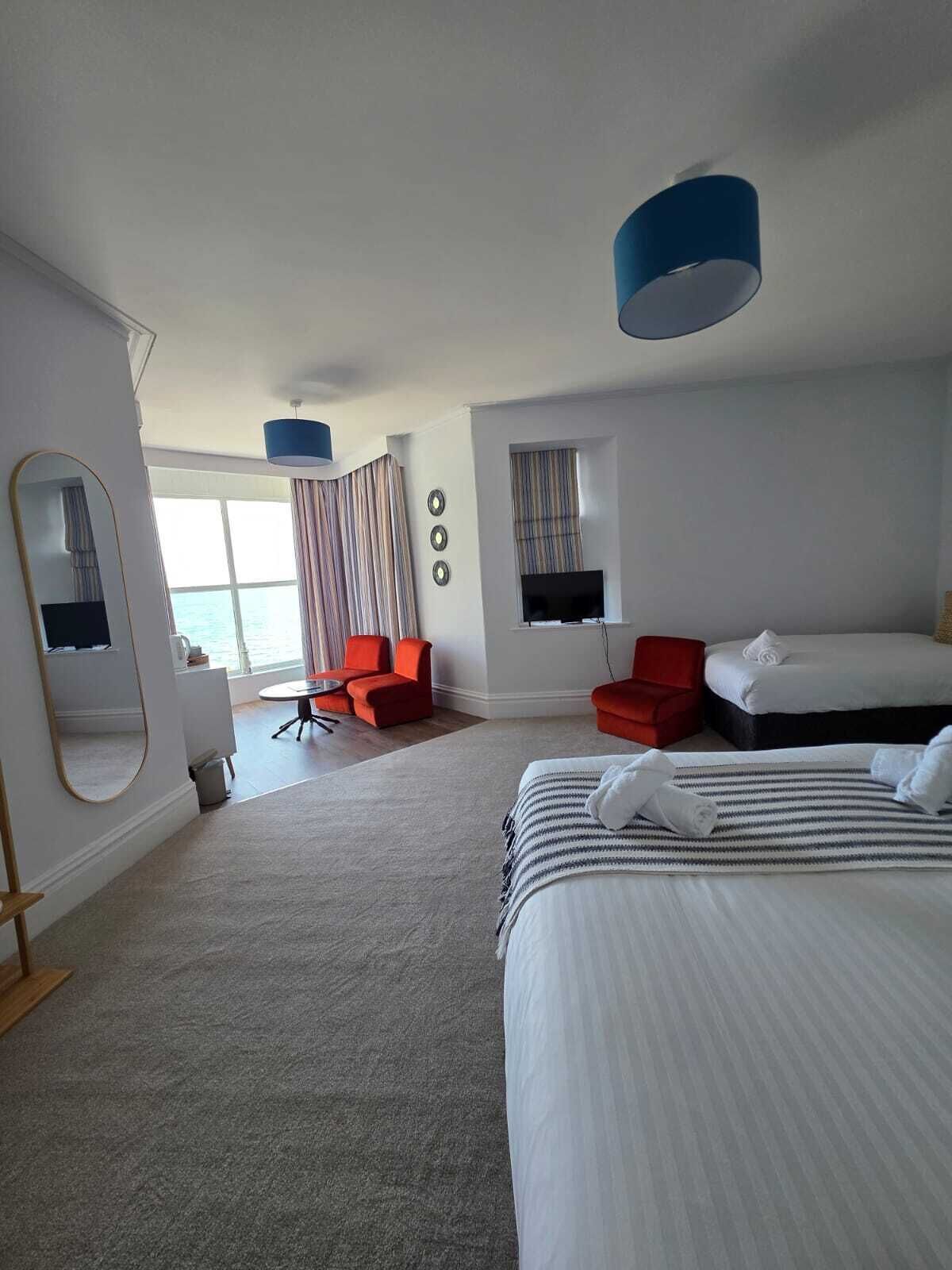 A hotel room with a large bed, a smaller bed, a desk, a chair, a TV, and a view of the sea.