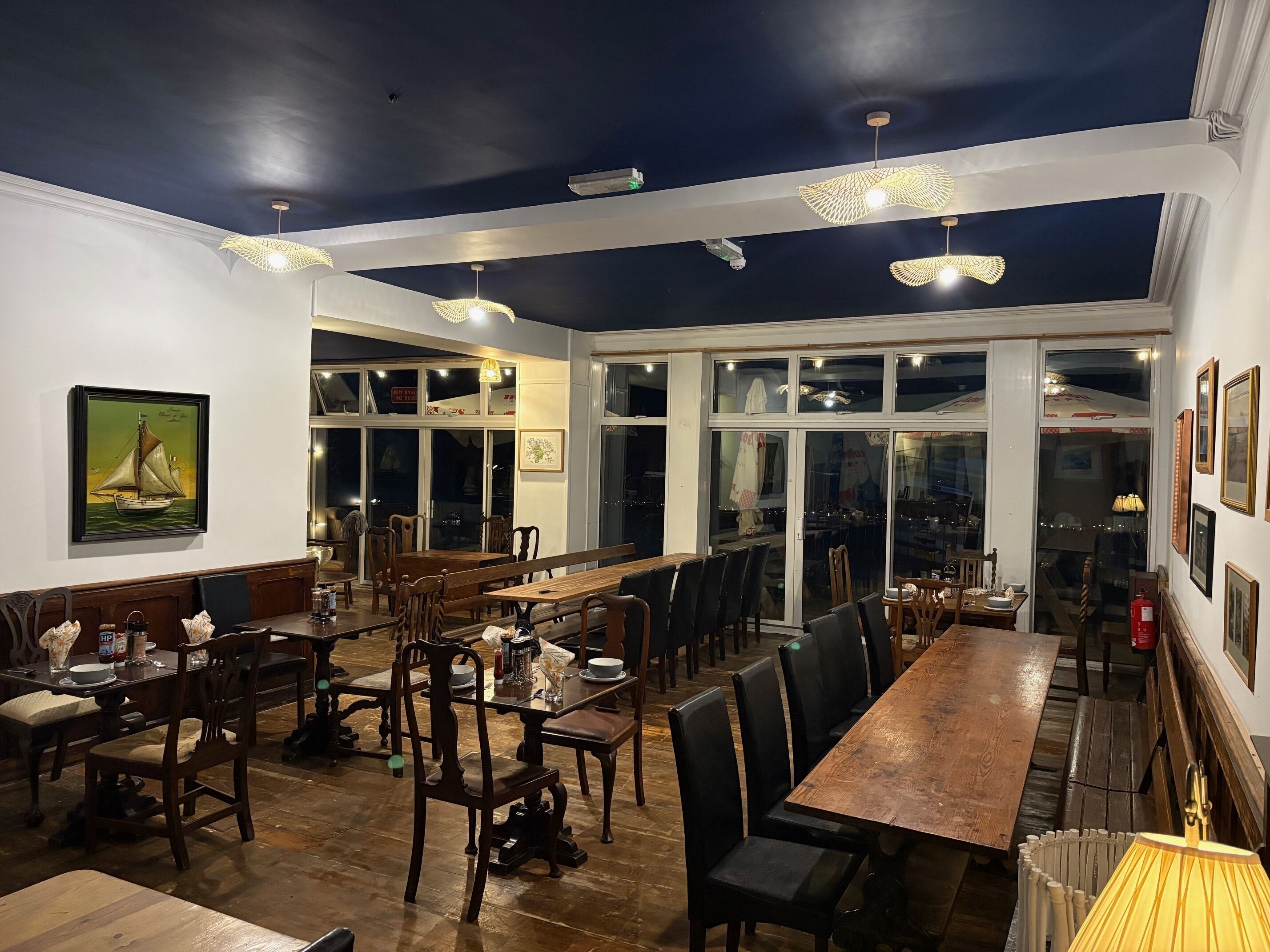 Uma sala de jantar ampla, com mesas e cadeiras de madeira, grandes janelas e luminárias pendentes.