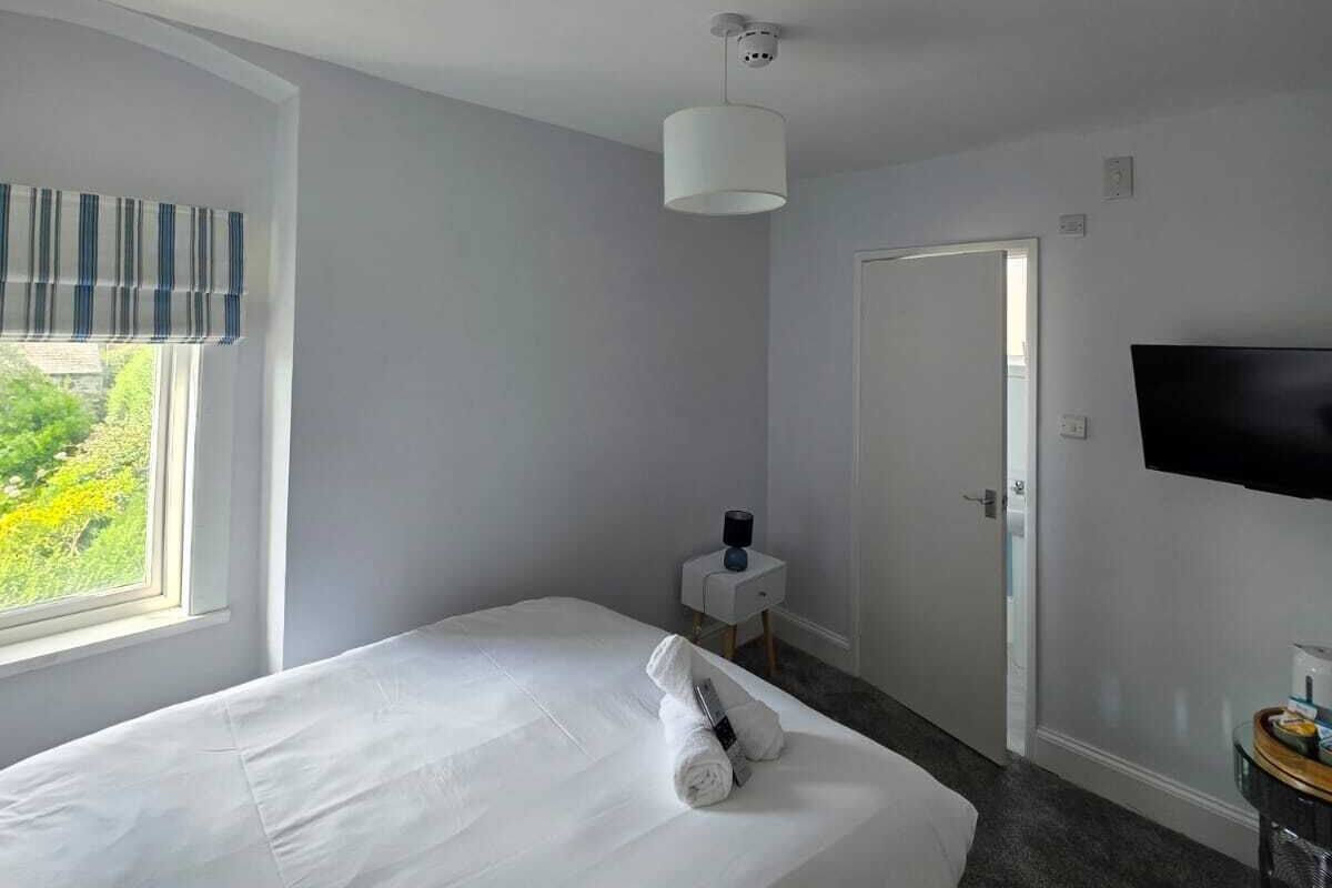 Ein Hotelzimmer mit Bett, Nachttisch mit Lampe, an der Wand montiertem Fernseher und Fenster mit Blick auf Grünflächen.