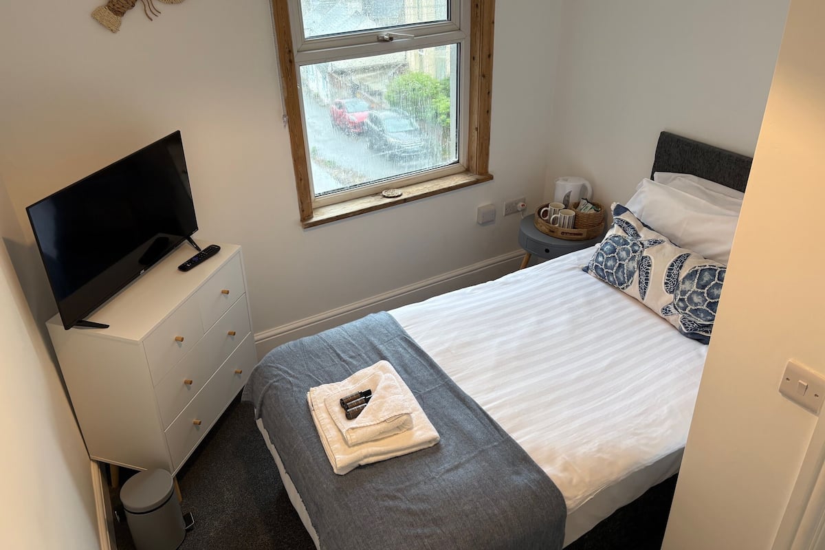 Ein Schlafzimmer mit einem Bett, einem Fernseher auf einer Kommode, einem Fenster mit Blick auf die Straße und einem Nachttisch mit Lampe und Korb.