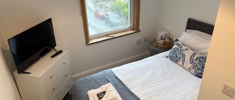 A bedroom with a bed, a TV on a dresser, a window with a view of the street, and a bedside table with a lamp and a basket.