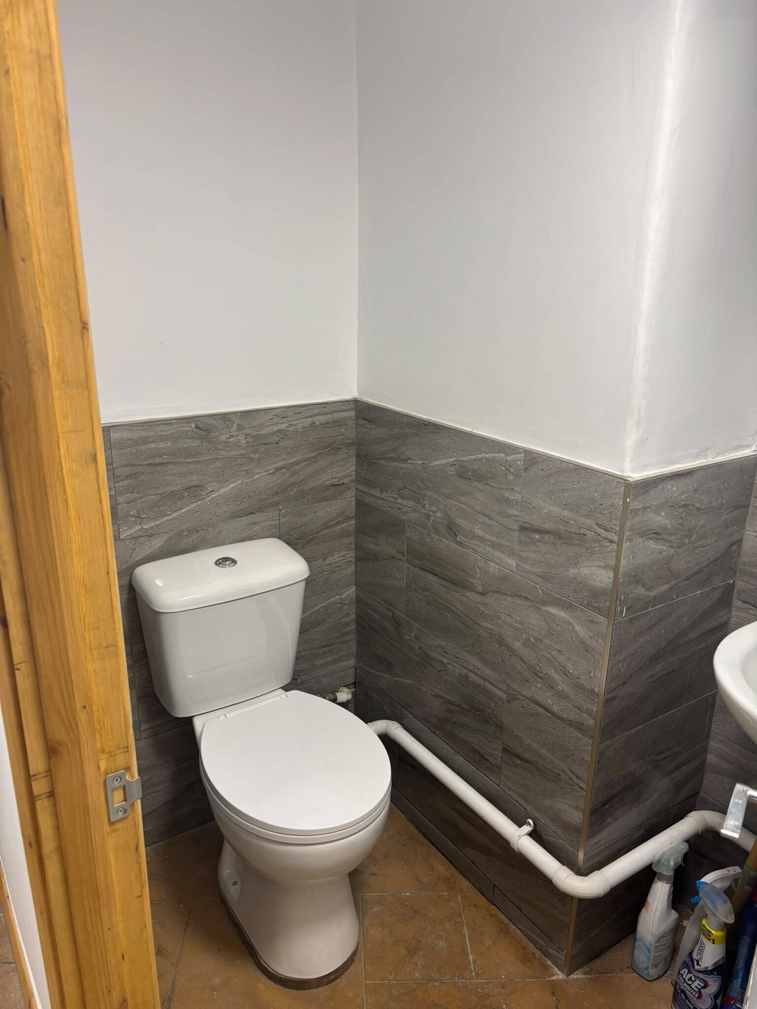 Une salle de bain avec des toilettes, des murs carrelés de gris et de blanc, et un cadre de porte en bois.