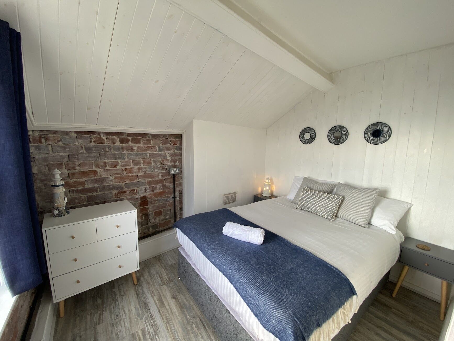A bedroom with a brick wall, a white dresser, a bed with a blue blanket, and three wall-mounted circular decorations.