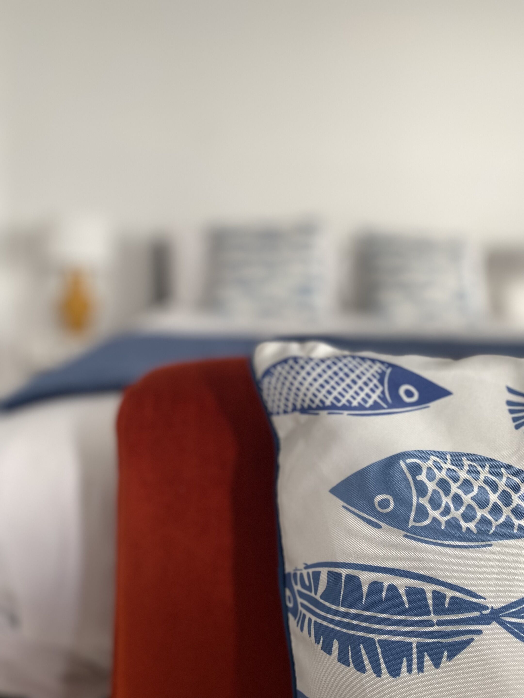 A bed with a blue and white pillow featuring fish patterns, a red pillow, and a white wall with a framed picture in the background.
