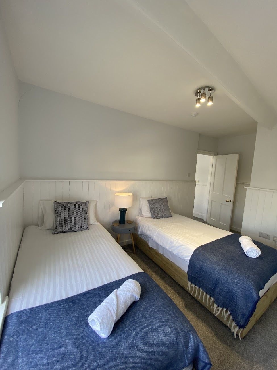 A hotel room with two beds, a wardrobe, a small table with a lamp, and a ceiling light fixture.