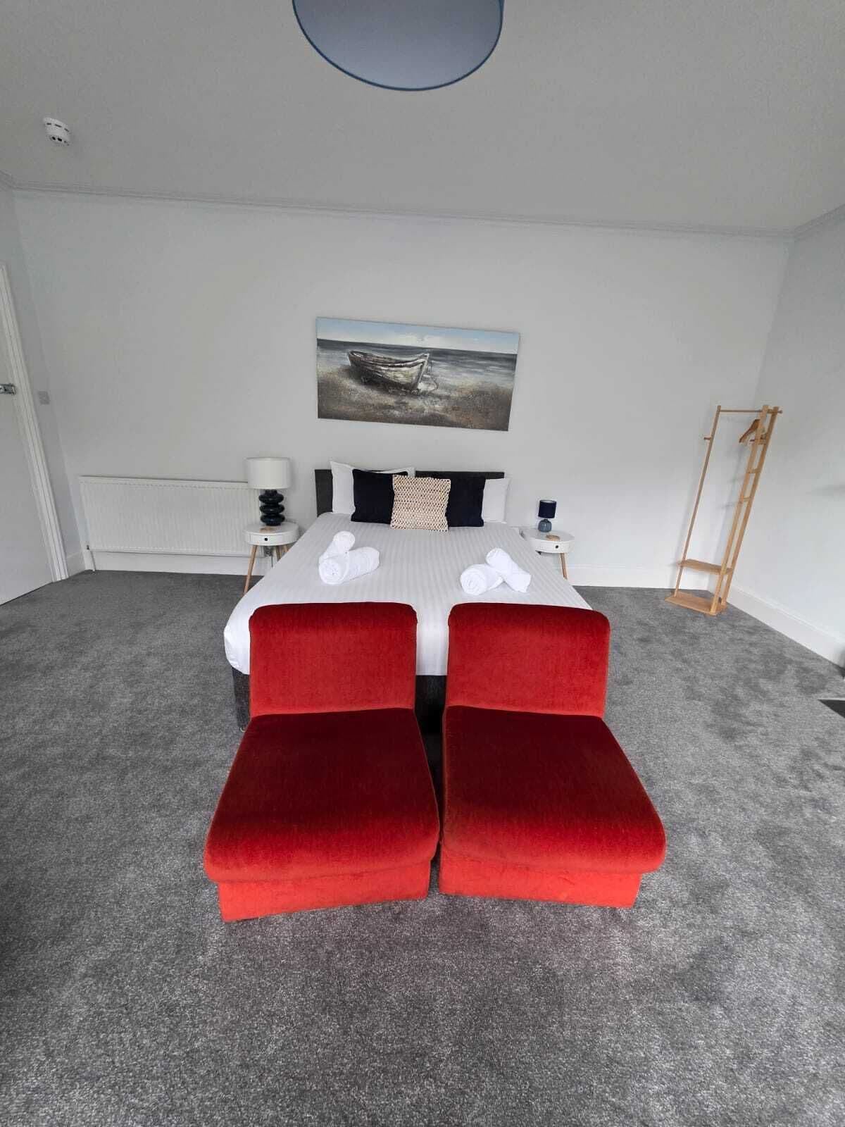 A bedroom with a bed, two red upholstered benches, a painting on the wall, a wooden ladder, and a white door.