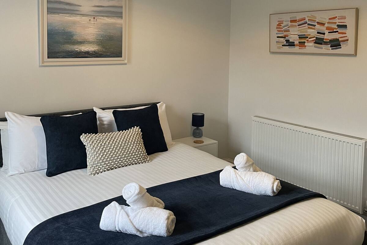 Ein ordentlich gemachtes Bett mit weißer und dunkelblauer Bettwäsche, einer schwarzen Tagesdecke und zwei Handtüchern. Darüber hängt ein gerahmtes Landschaftsbild, an der Wand hängt ein farbenfrohes abstraktes Gemälde.