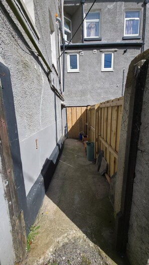 Un callejón estrecho con piso de concreto, cerca de madera y un edificio con varias ventanas y una puerta.