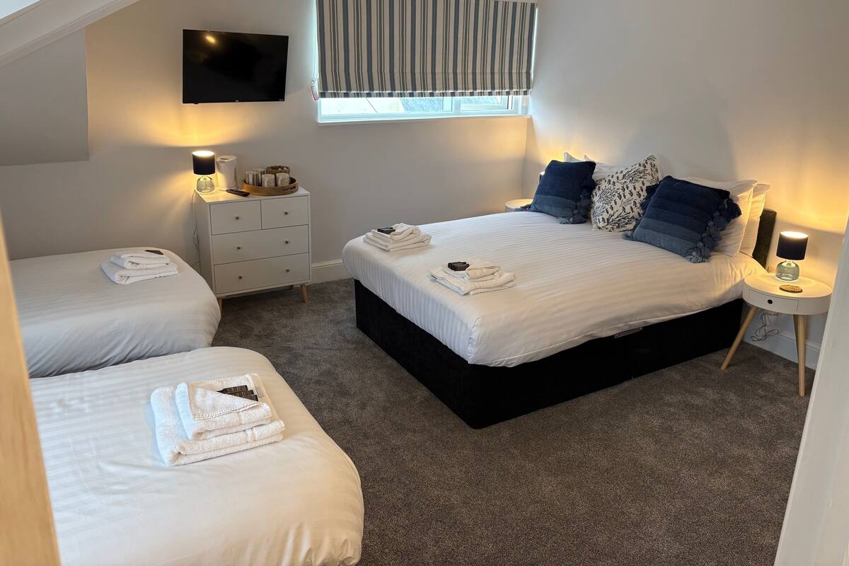 Ein Schlafzimmer mit einem großen Bett, zwei Einzelbetten, einem Fernseher, Nachttischen und einem Fenster mit Vorhängen.