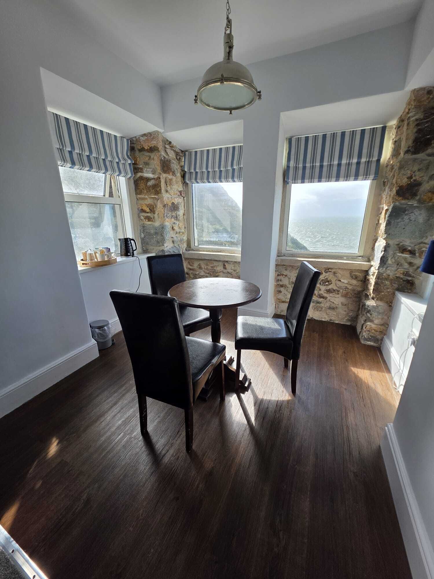 A dining area with a round table and four chairs, stone walls, large windows with striped curtains, and a hanging light fixture.