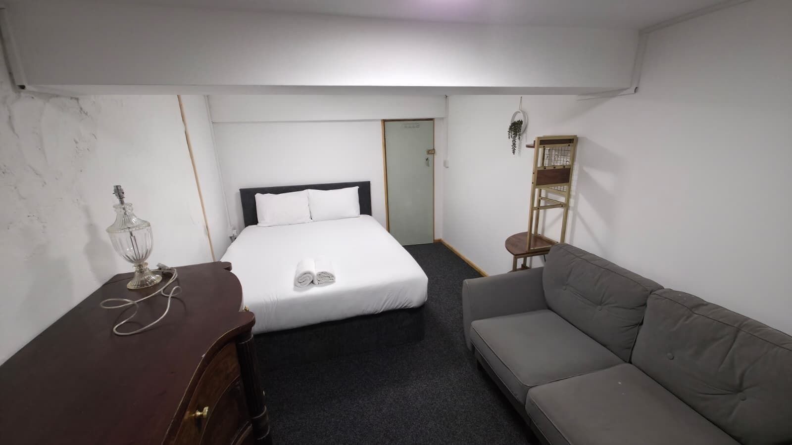 A compact hotel room with a bed, a sofa, a desk with a lamp, and a small plant.