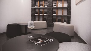 A modern interior with a round table, a glass of wine, and a magazine. There are cushioned seating areas and a wall with shelves of bottles. - Weingut Gehlen-Cornelius (Brauneberg)