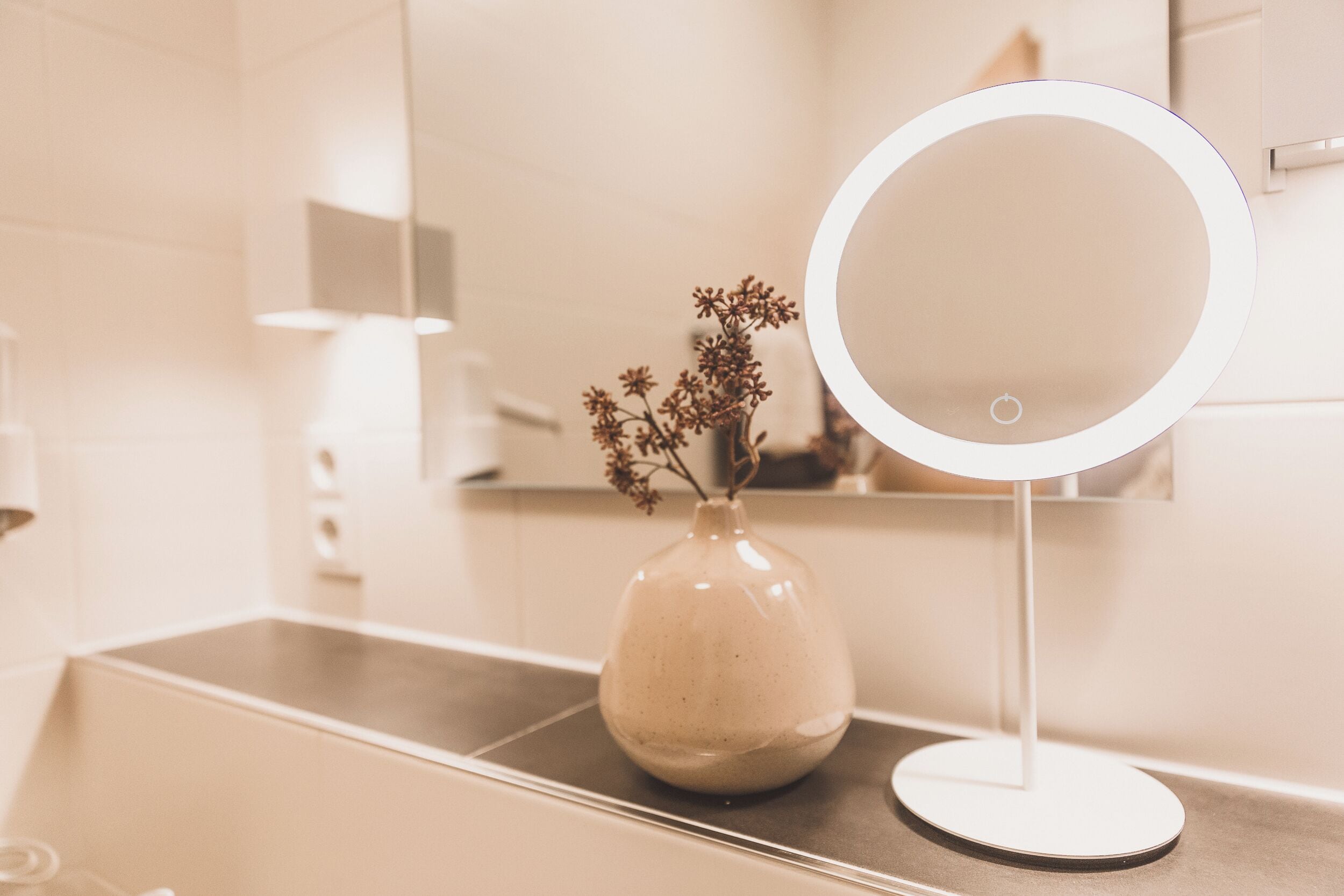 A bathroom vanity with a round mirror, a vase with dried flowers, and a modern light fixture.