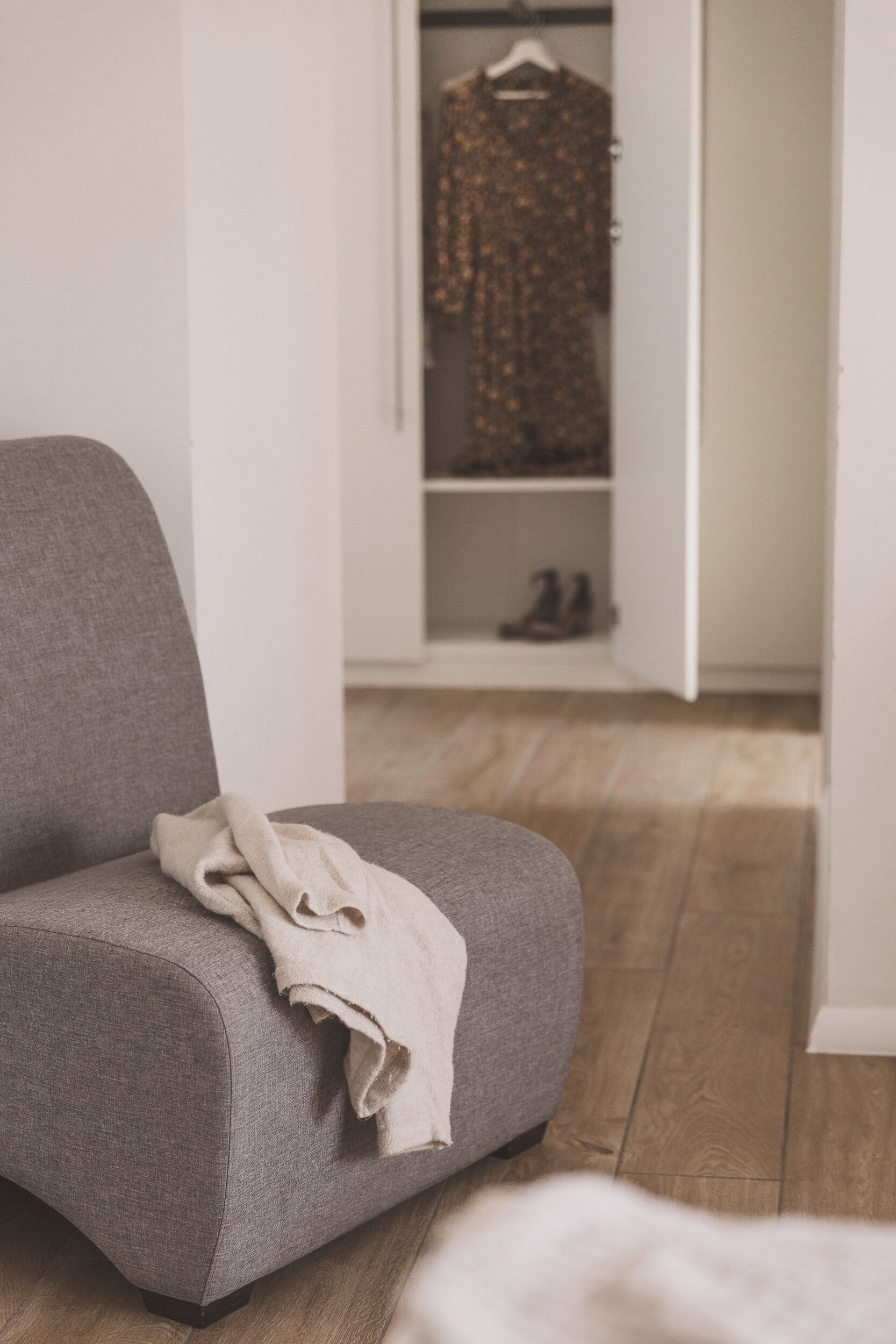 A grey armchair with a beige throw blanket, a wooden floor, and a closet with a hanging garment and shoes.