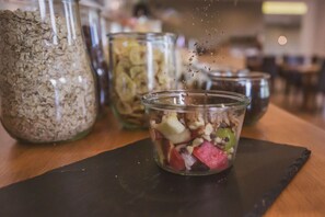 A clear glass with a mix of fruits and nuts on a wooden surface, with a jar of oats and a blurred background. - Weingut Gehlen-Cornelius (Brauneberg)