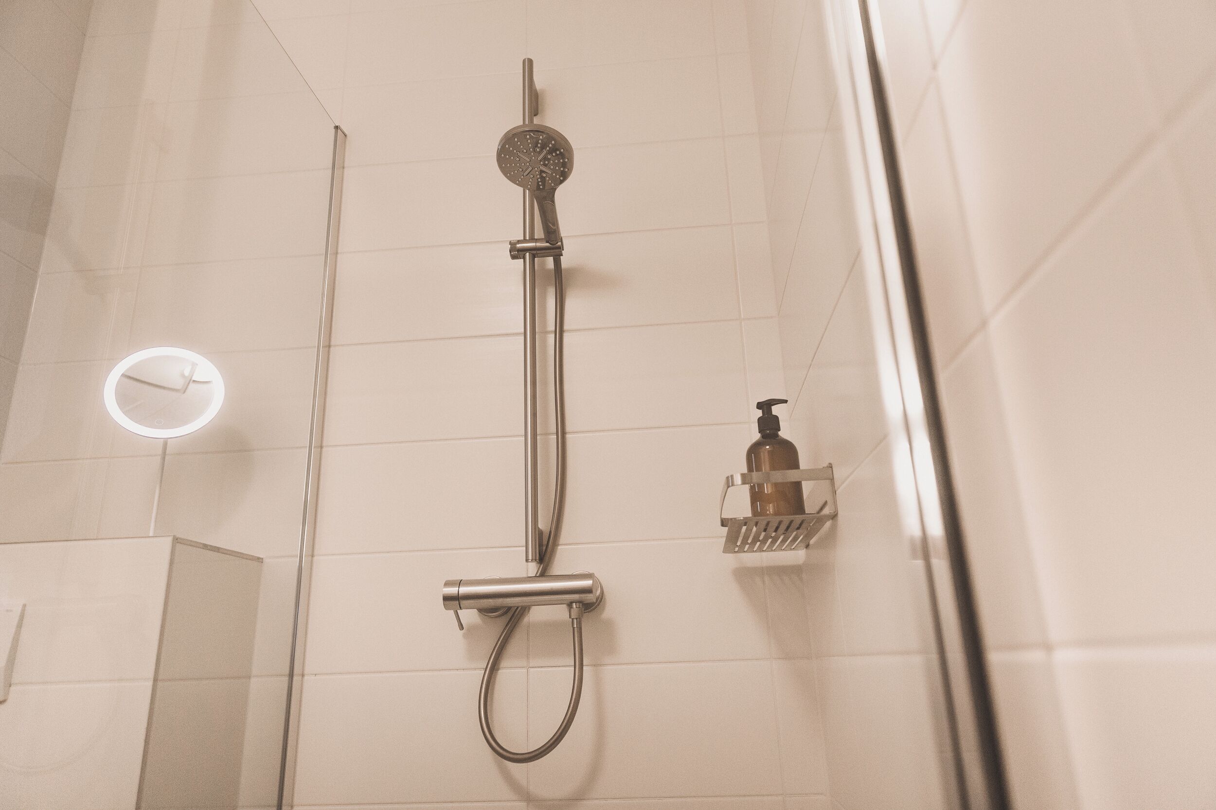 A modern bathroom with a glass-enclosed shower, a round mirror, and a soap dispenser on a shelf.