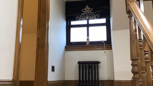 Un escalier avec une rampe en bois mène à un palier. On peut apercevoir un radiateur sous une fenêtre ornée d’une lambrequin décorative.