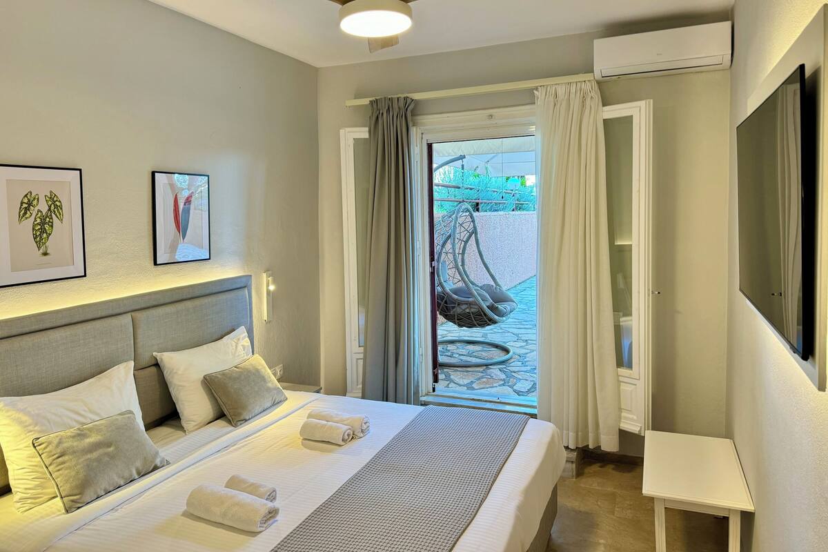 A hotel room with a bed, a ceiling fan, a TV, and a view of the sea through a glass door.