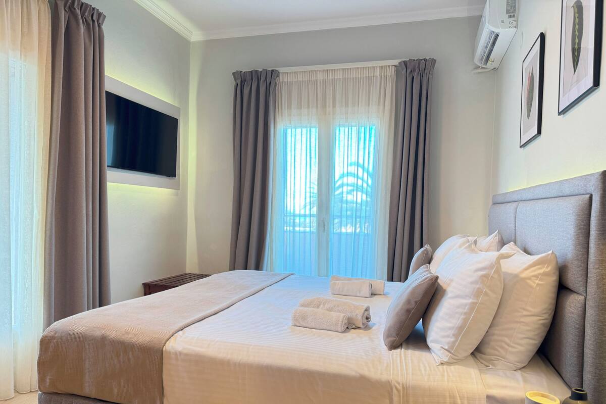 A neatly made bed with a headboard, a bedside table with a remote control, and a window with curtains.