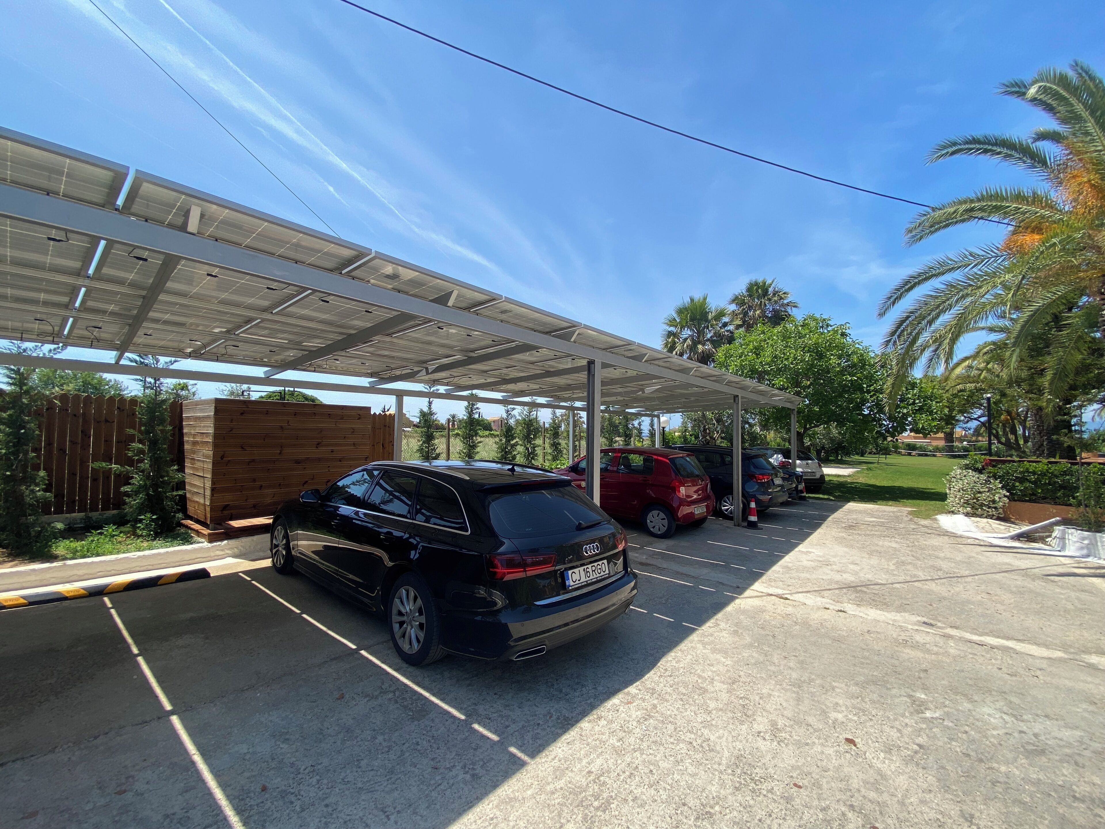 A covered parking area with cars parked under a roof, surrounded by greenery and palm trees.