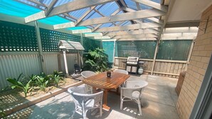 Un patio con mesa de madera, sillas de mimbre, una parrilla y valla verde.
