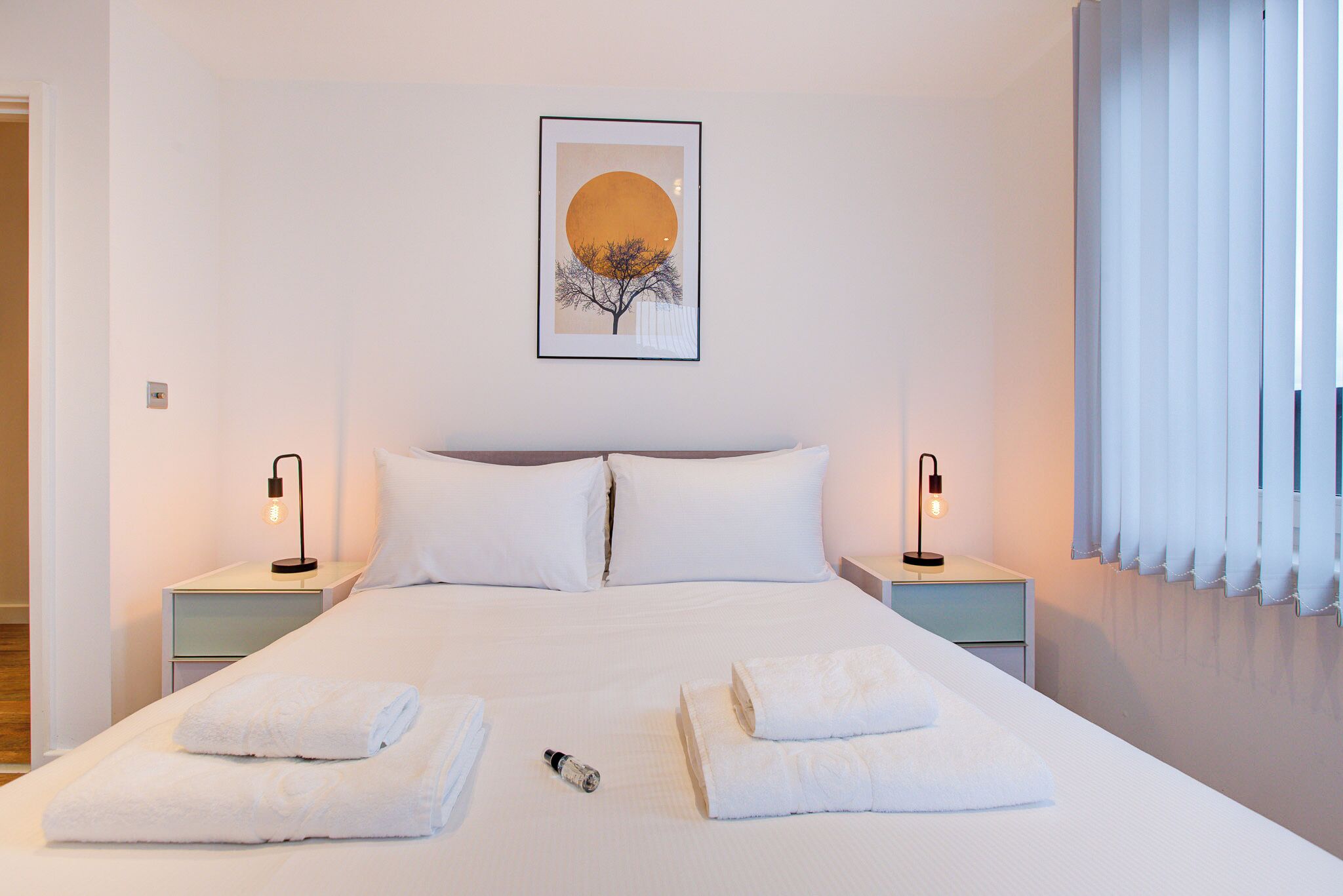 A neatly made bed with white linens and towels, two bedside tables with lamps, and a framed artwork above the bed.