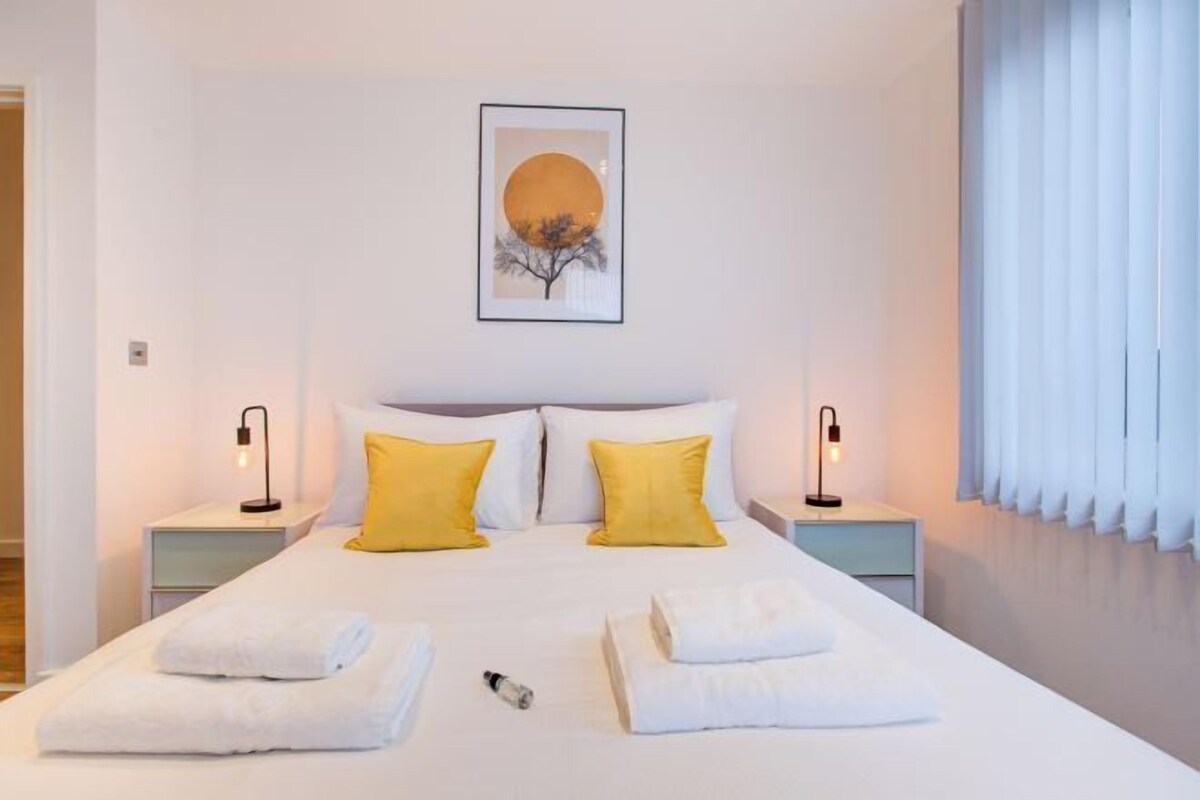 A neatly made bed with white and yellow pillows, a framed artwork above, and a window with blinds.