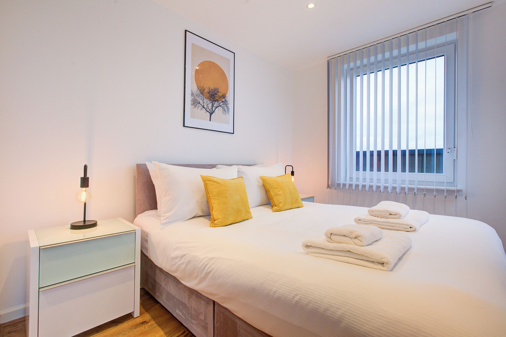 A neatly made bed with white and yellow pillows, a bedside table with a lamp, and a window with blinds.
