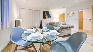 A modern living room with a glass dining table, blue chairs, a sofa, and a painting on the wall. - Hybrid Resi Greenwich (London)