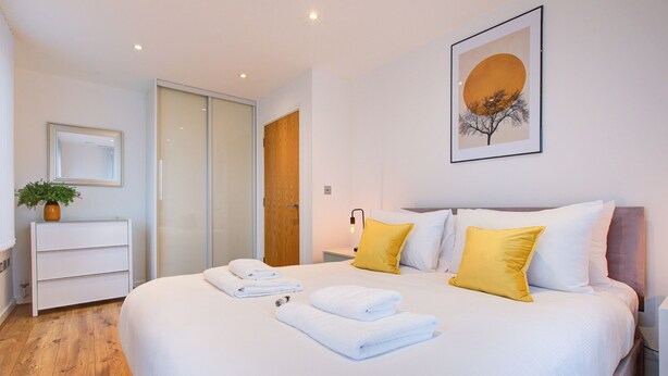 A hotel room with a large bed, white bedding, a wooden floor, a bedside table with a lamp, a mirror, and a framed picture on the wall. - Hybrid Resi Greenwich (London)
