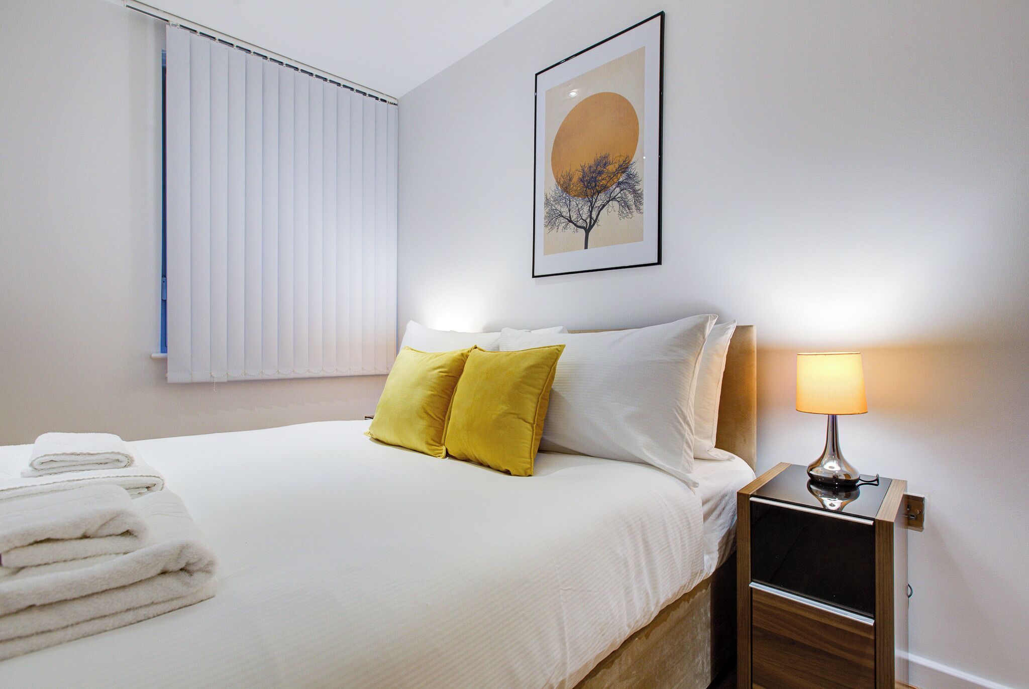Ein ordentlich bezogenes Bett mit weißen und gelben Kissen, ein gefaltetes weißes Handtuch und ein Nachttisch mit einer Lampe.