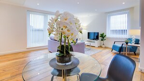 A modern living room with a glass dining table, a sofa, a TV, and a potted orchid plant. - Hybrid Resi Greenwich (London)