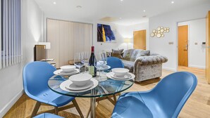 A modern living room with a glass dining table, blue chairs, a sofa, and a painting on the wall. - Hybrid Resi Greenwich (London)