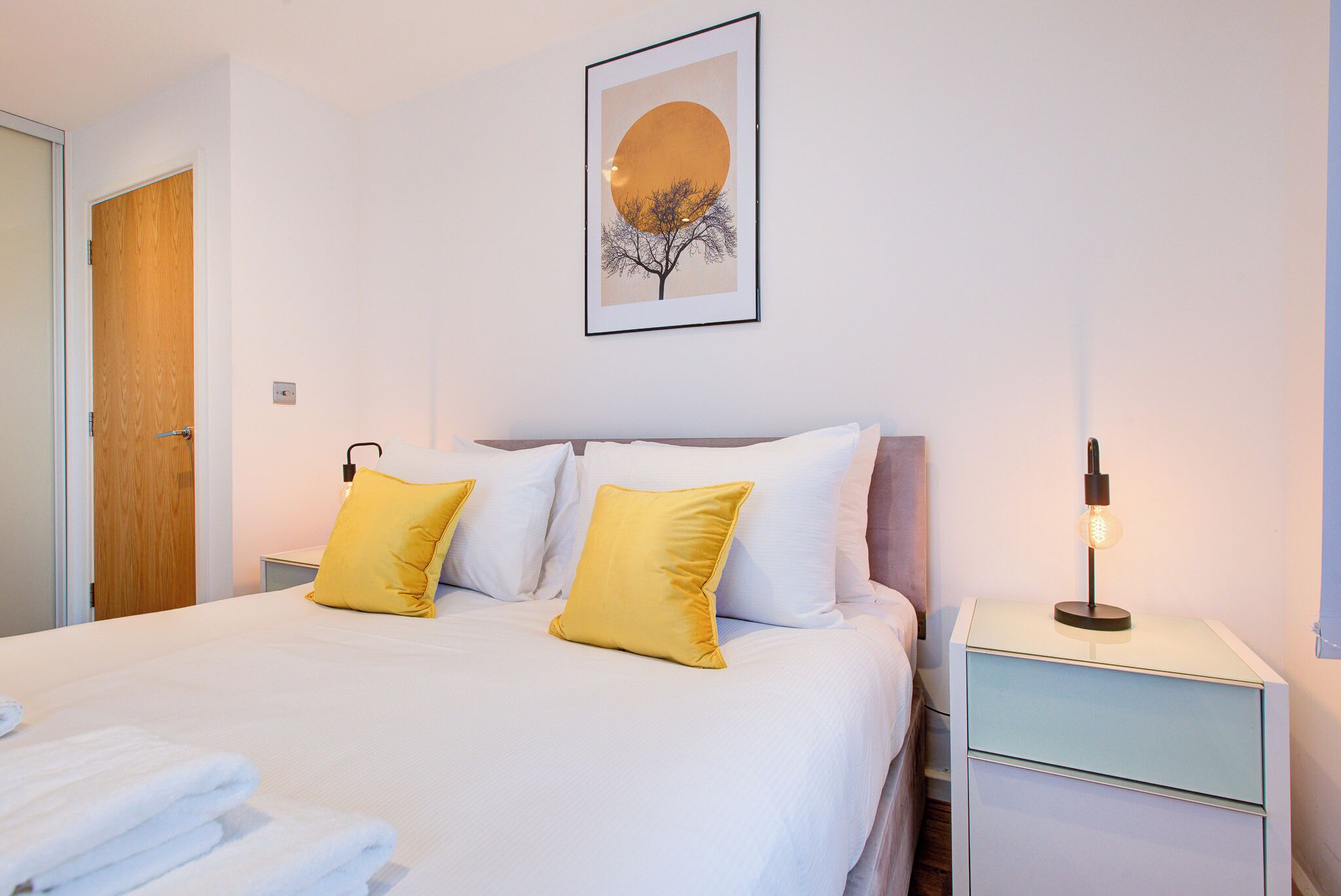 A neatly made bed with white and yellow pillows, a framed picture on the wall, and a bedside lamp.