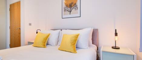 A neatly made bed with white and yellow pillows, a framed picture on the wall, and a bedside lamp.