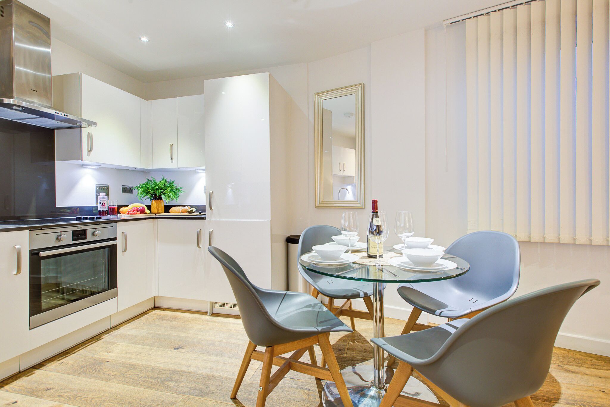 A modern kitchen with white cabinets, a stainless steel range hood, and a built-in oven. There is a dining table with four chairs and a glass top.