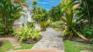 A pathway leading through a lush garden with various tropical plants and a white building in the background. - Turtle View by Bluescape (Holetown)
