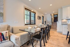 Uma sala de jantar com uma mesa comprida posta para seis pessoas, com um sofá e uma escada que leva a uma cozinha.