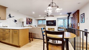 A modern kitchen with wooden cabinets, a dining area with a table and chairs, a sofa, and a TV mounted on the wall.