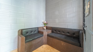 A corner seating area with two black leather sofas, a small wooden table with a vase of flowers, and a white brick wall. - Seasons Suites HBR (Bengaluru)