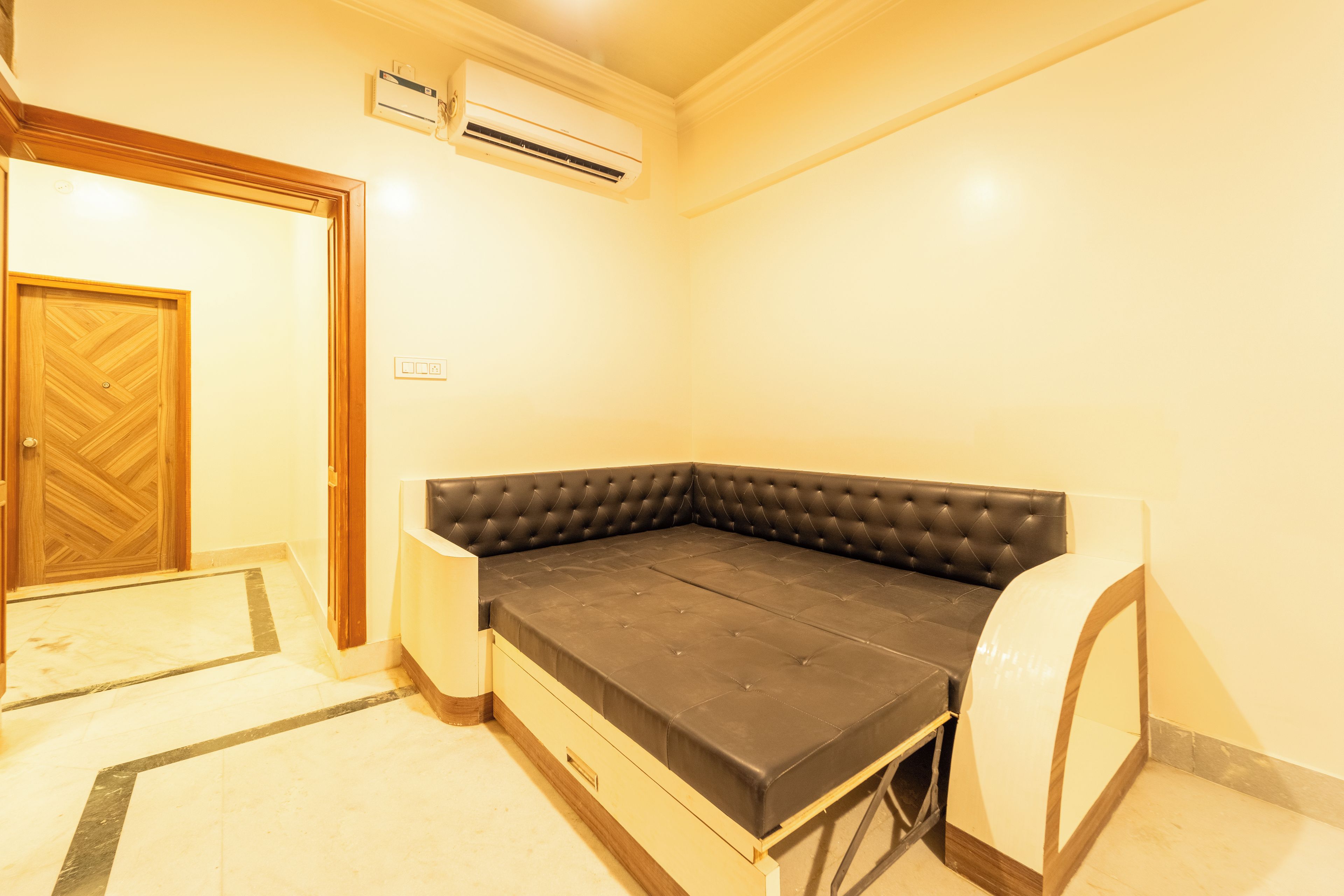 A room with a corner sofa, an air conditioning unit, a wooden door, and a small table.