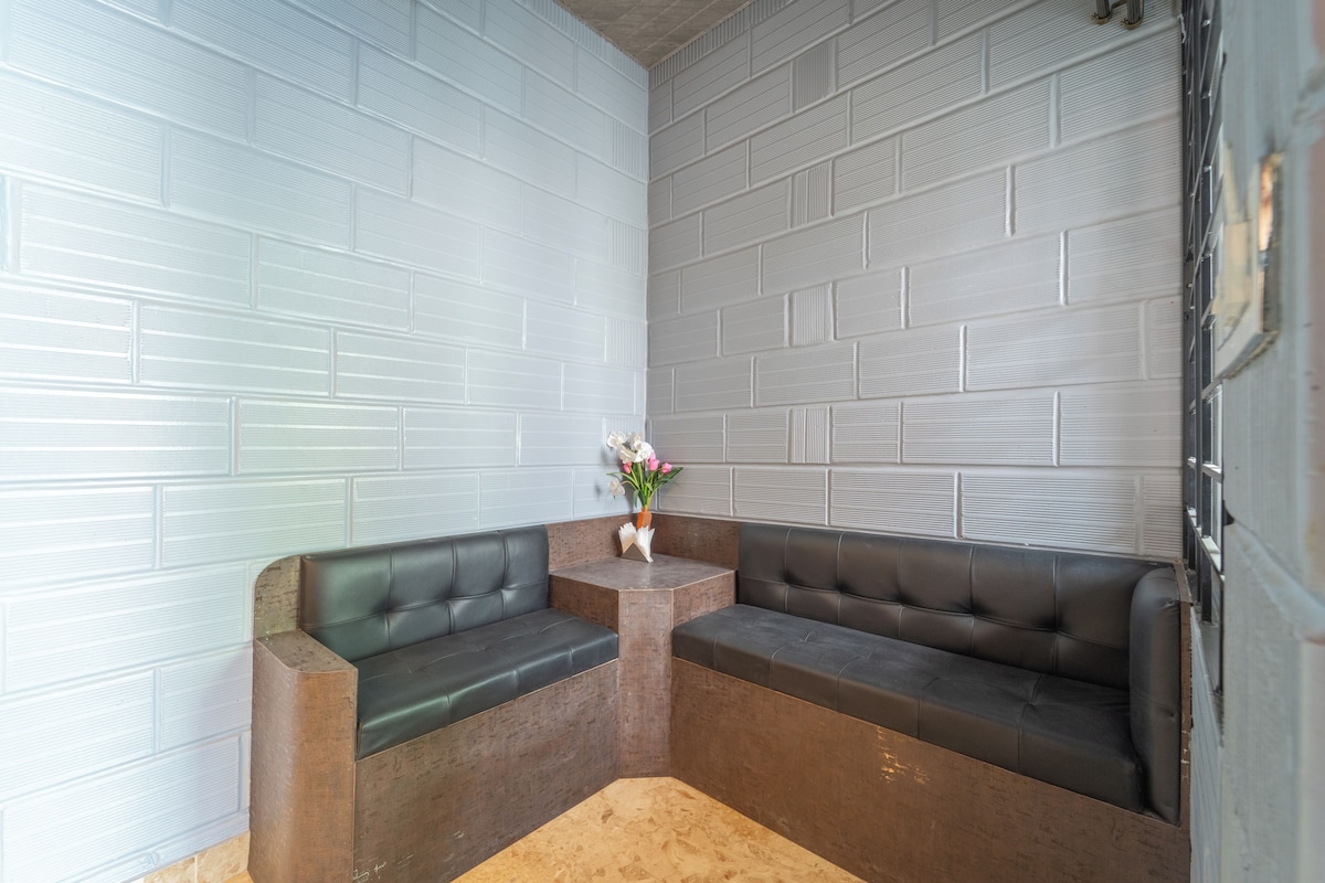 Un rincón con asientos que incluye dos sofás de cuero negro, una mesita de madera con un florero y una pared de ladrillo blanco.