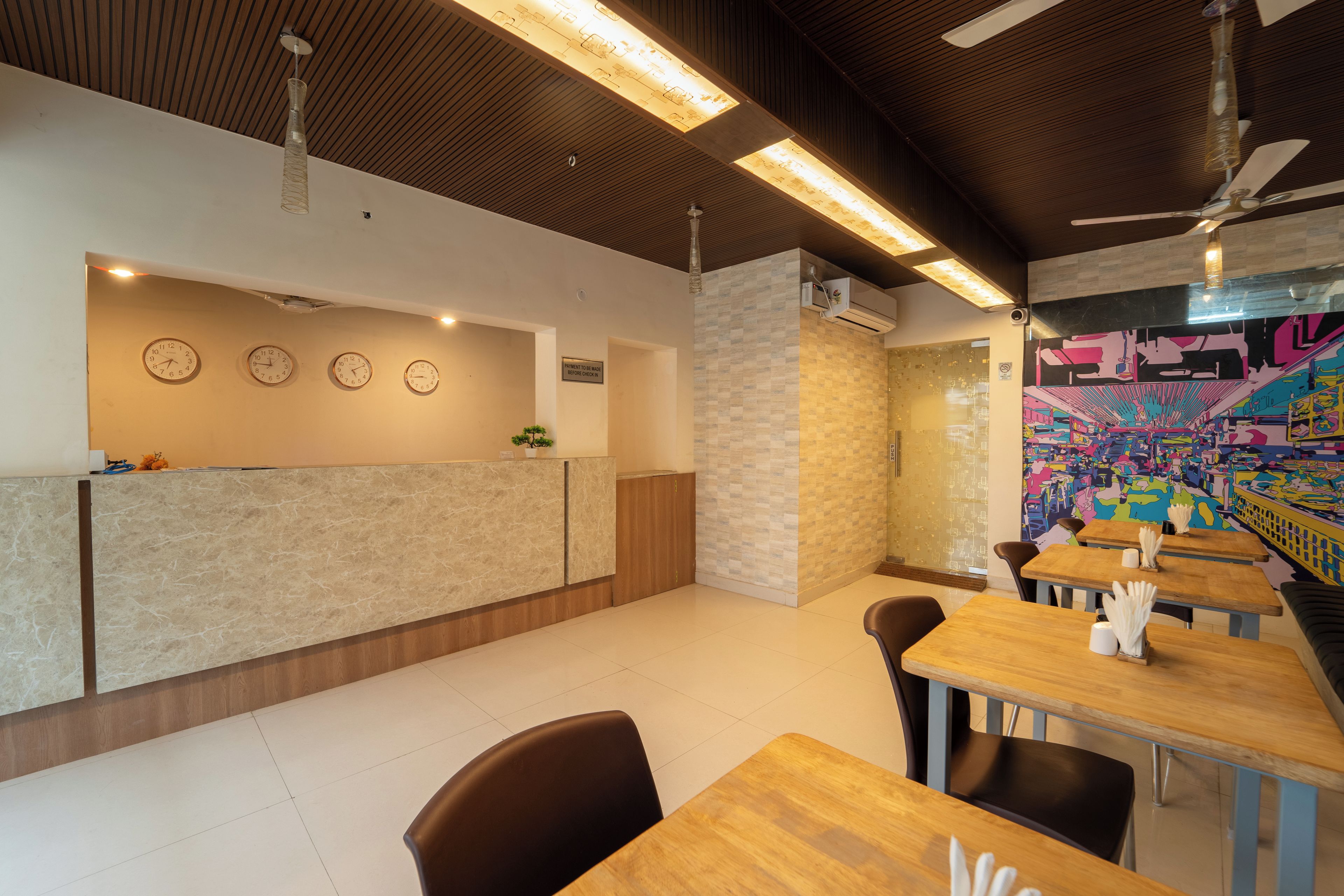 Interior de restaurante con mostrador de recepción, relojes en la pared, ventilador de techo y un mural colorido.