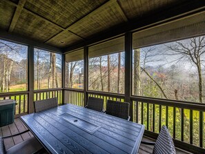 A covered outdoor area with a wooden table and chairs, large windows offering views of a forested landscape. - Spacious Lake Toxaway Home with Golf Course Views (Lake Toxaway)